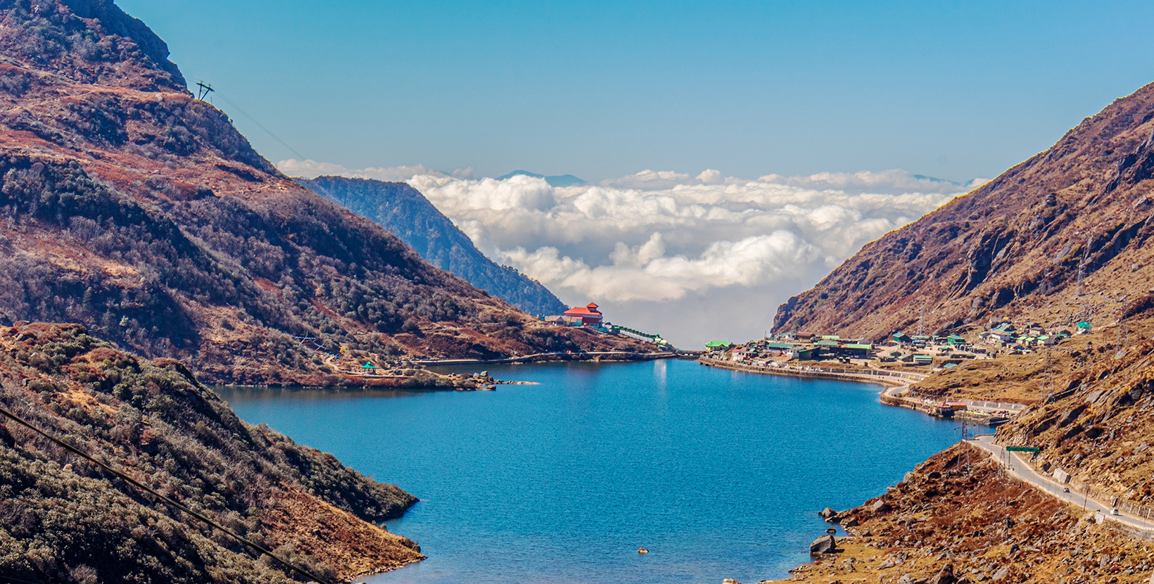 A blue water Tsomgo lake surrounded by desert brown mountains is located high above clouds. The winding road to the distant village leads a way to heaven in cumulus clouds and blue sky. Sikkim, India